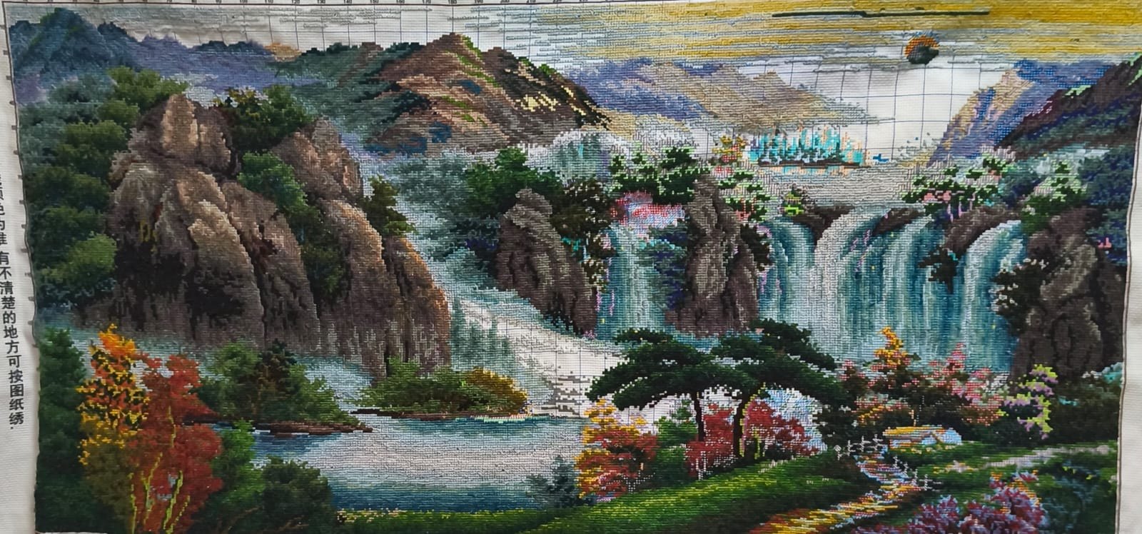 Hand-stitched cross-stitch of a mountain landscape with cascading waterfalls, a blue lake, autumn trees and misty peaks