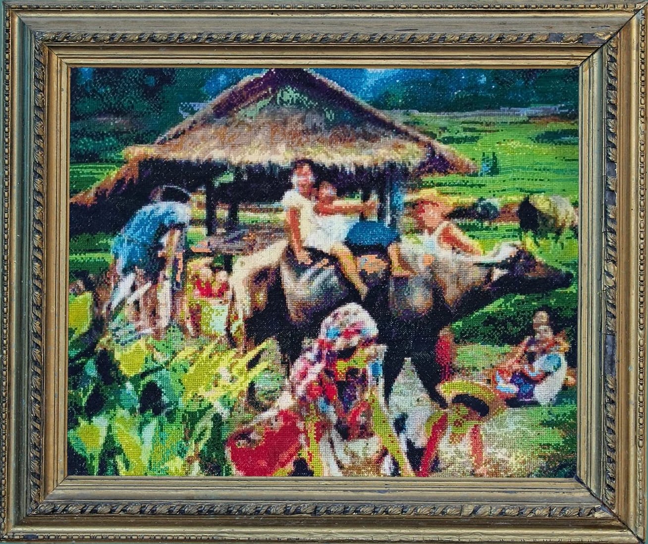 Hand-stitched cross-stitch of a rural barrio scene: a thatched nipa hut, villagers and children gathered around a water buffalo, with green rice fields behind