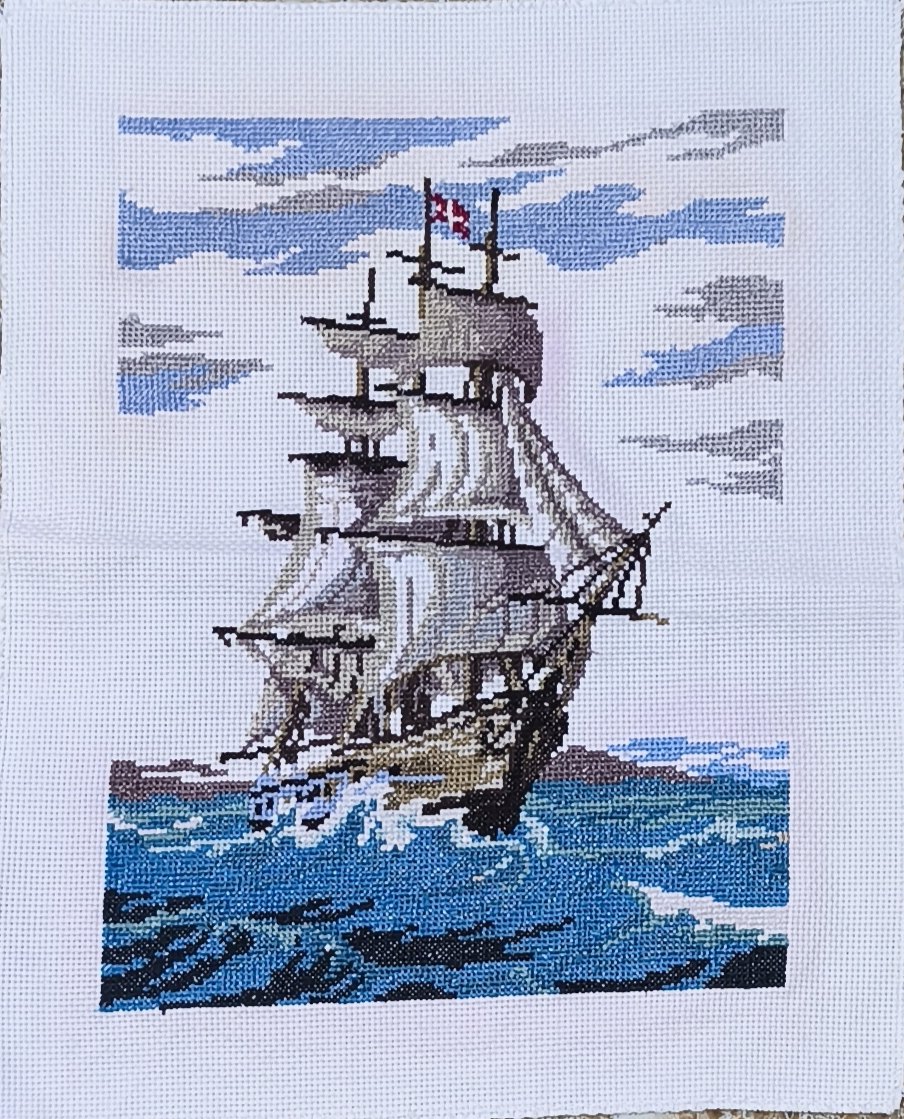 Hand-stitched cross-stitch of a square-rigged sailing ship under full sail on a rough blue-green sea, beneath a sky of blue and grey cloud, with a small red and white flag at the masthead